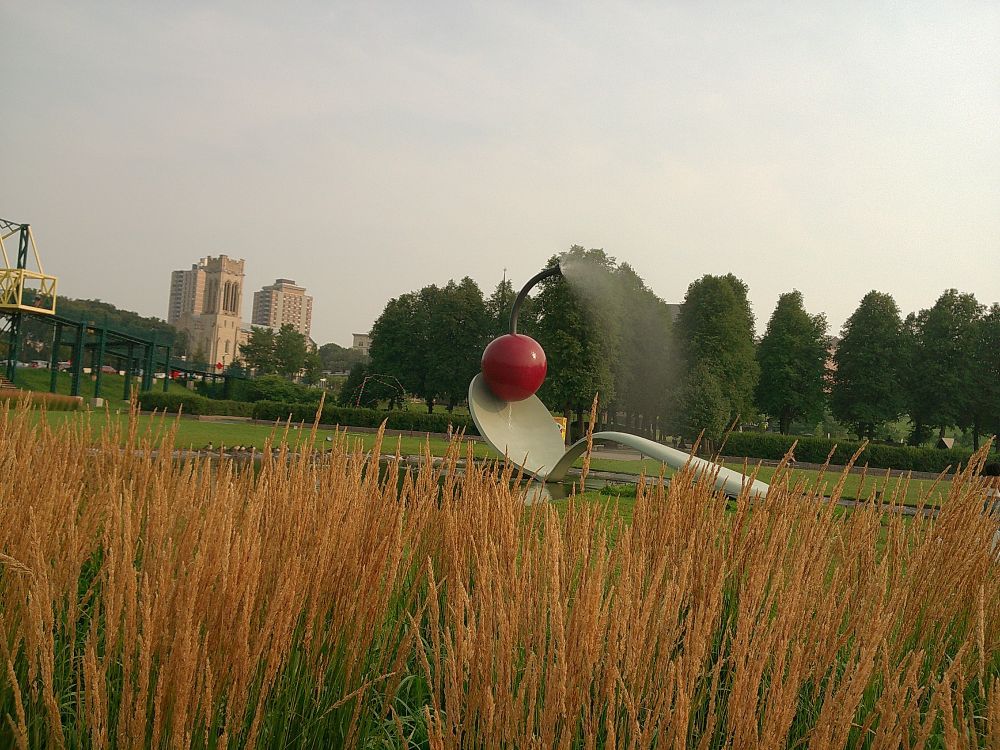 photo of a large sculpture outside of a spoon with a red cherry on top that is spraying water out of the stem