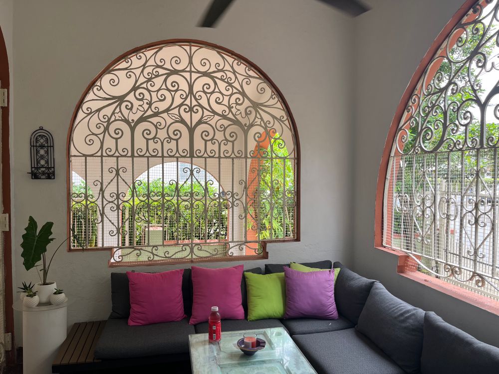 A porch with a dark gray couch and bright pink, green, & purple pillows. The big windows are arched and filled in with fancy curved grillwork. Through them you can see more arches and greenery