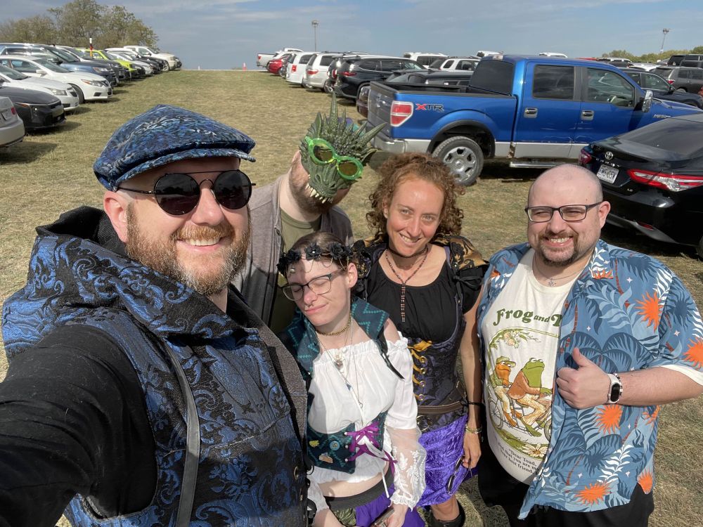 A group of people prepare to enter the ren fest. From left to right: a tall man with a preying beard wears a vest that looks like an old timey blue and black sofa, a flat cap, and aviators. A man in a dragon mask and sunglasses. A tiny woman in a peasant blouse and corset closes her eyes to the sun. A taller red headed woman, hair very curly, wears a different peasant blouse and corset. And finally WT, a chubby bald man with glasses and a beard. He wears a frog and toad t-shirt and a blue and orange Hawaiian outer shirt. 