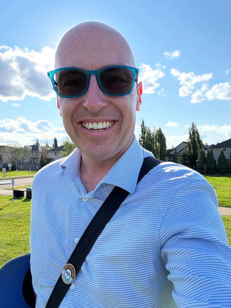Andrew wearing a blue and white shirt standing outside under a blue sky with some clouds.