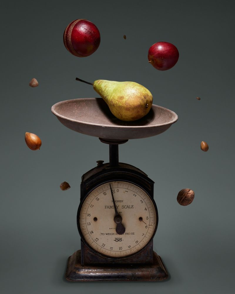 Eine alte Küchenwaage im Mittelpunkt. Darum herum fliegt Obst. 
