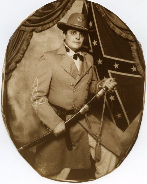 An "olde tyme" sepia photo of a man in a light colored outfit reminiscent of a Confederate officer's, posed standing in front of a Confederate Battle Flag