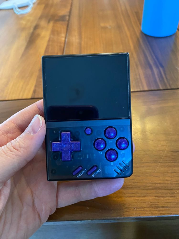 Hand holding a small black upright handheld game console with a purple d pad and purple buttons, set in front of a wooden table.