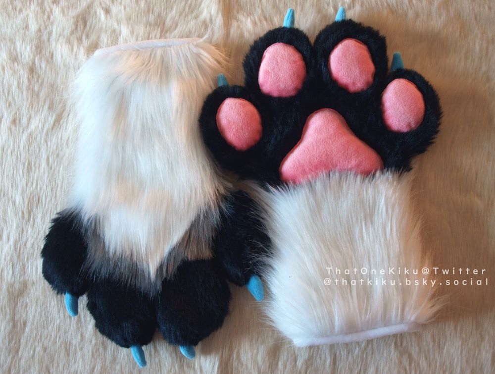 A pair of fursuit handpaws. White arm, black fingers and palm, a grey line over the knuckles, pink paw pads, blue claws.