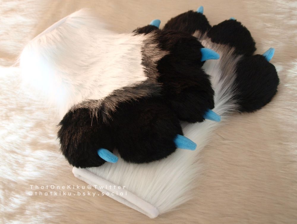 A pair of fursuit handpaws. White arm, black fingers and palm, a grey line over the knuckles, pink paw pads, blue claws.