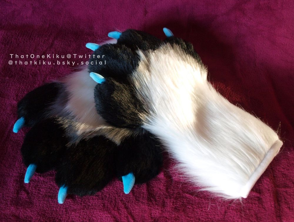 A pair of fursuit handpaws. White arm, black fingers and palm, a grey line over the knuckles, pink paw pads, blue claws.