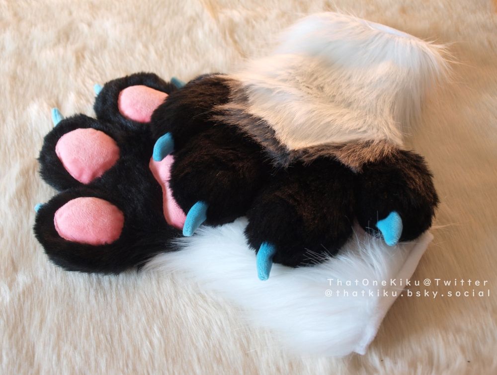 A pair of fursuit handpaws. White arm, black fingers and palm, a grey line over the knuckles, pink paw pads, blue claws.