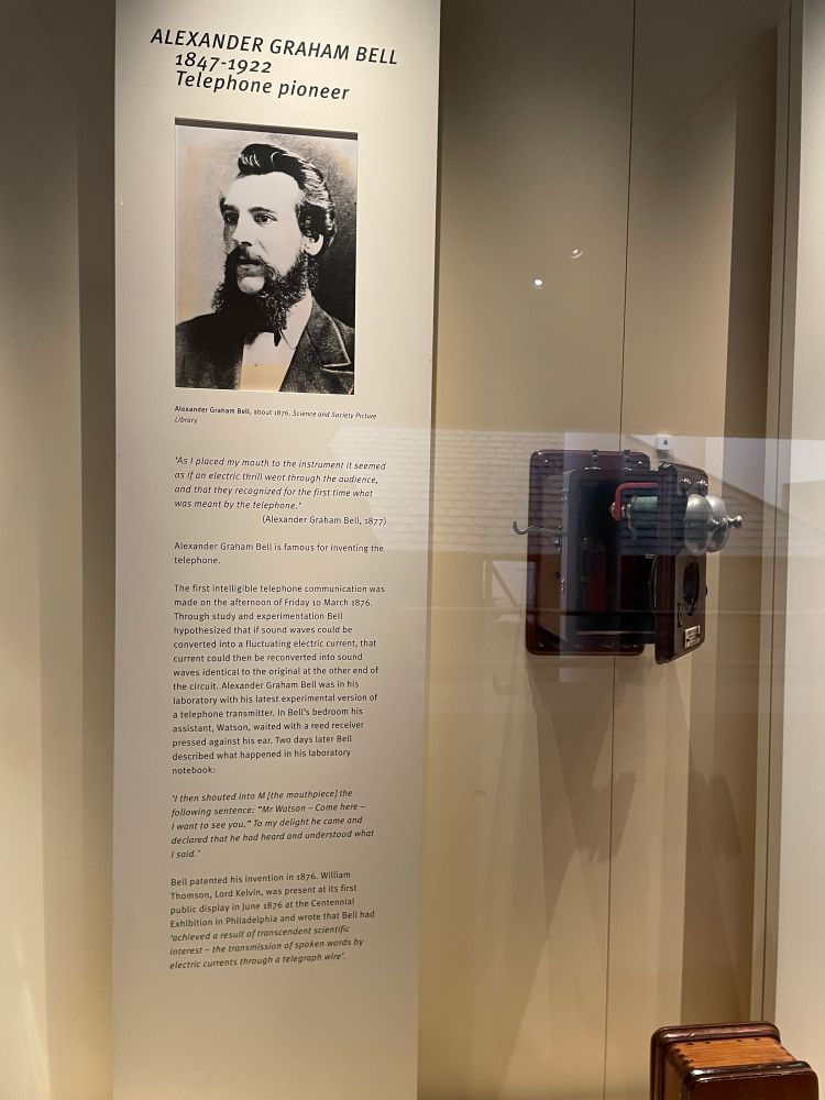 Hinter einem Glasfenster in einem Museum sieht man links eine Tafel und rechts ein Telefon. Auf der Tafel steht: Alexander Graham Bell 1847-1922 Telephone pioneer. Darunter ein Foto von Bell und ein Text zum ersten Versuch eines Telefonats.