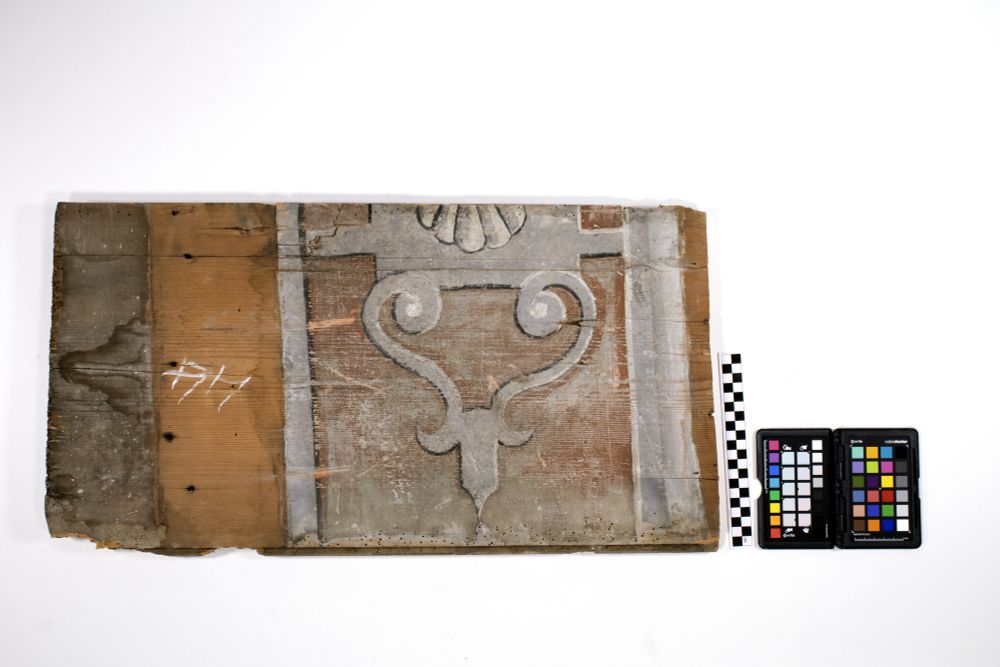 Photograph of a loose ceiling board dating from 1581, with a colour checker and ruler. The ceiling board is part of the Prestongrange House ceiling which is nowadays housed in Edinburgh Napier University's medieval Merchiston Tower. The board in question features - in shades of black, white, and gray - part of a shell (scallops) and swirls resembling ornate ironwork, on a background of red.