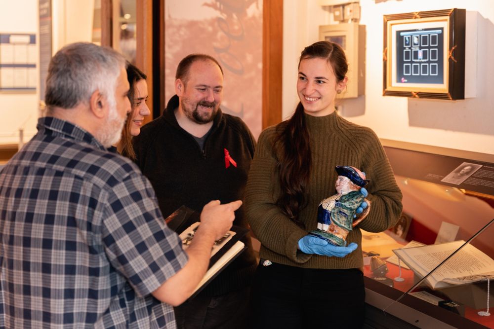 Photograph of ENU Curator showing a toby jug to three other people during a tour in the War Poets Collection exhibition at Edinburgh Napier University's Craiglockhart Campus, which was a hospital for shell-shocked officers during WWI. The toby jug (a jug in the shape of a standing man with a green coat, yellow waistcoat, blue trousers, white stockings, and a blue wide-brimmed hat, was hand painted by Second Lieutenant J. Wood as part of his treatment at the hospital in 1918. 