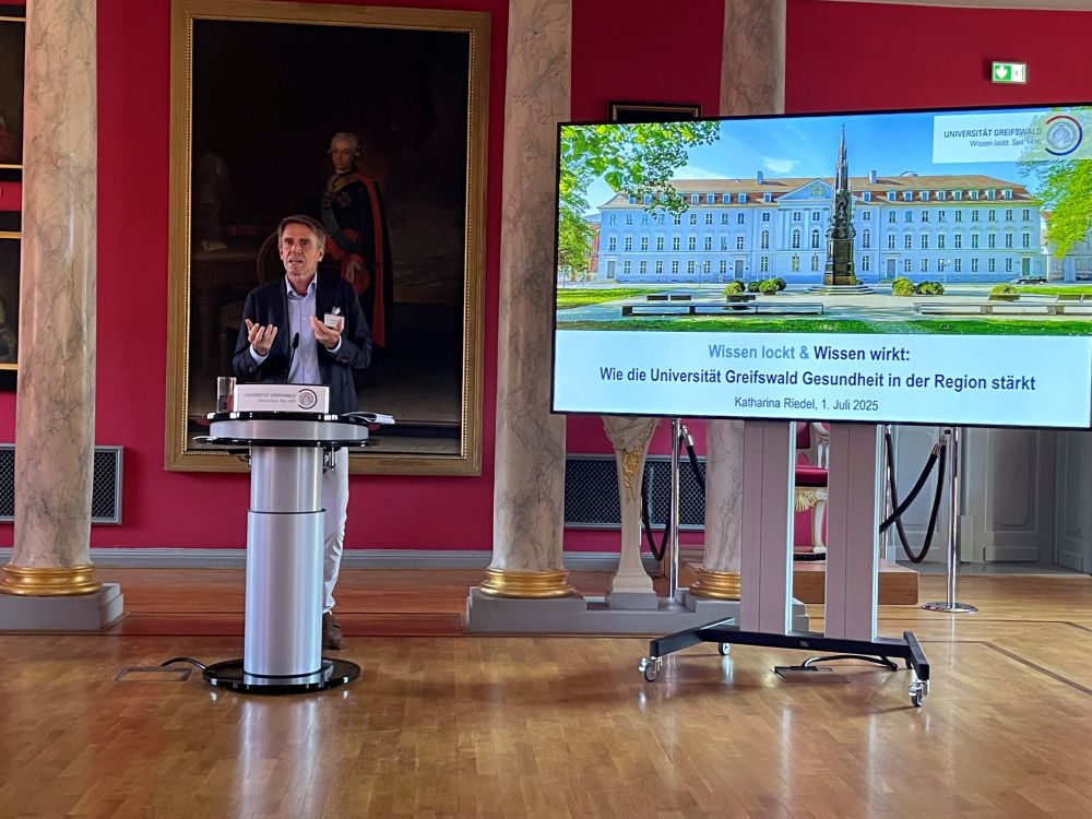 Wolfgang Wick beim Vortrag zur Gesundheitsförderung in einem ehrwürdigen alten und aufwändig sanierten Raum der Uni Greifswald. Im Hintergrund ein riesiges altes Gemälde mit goldenem Rahmen auf einer weinrot gestrichenen Wand. Außerdem: Marmorsäulen mit goldenem Sockel. Wick steht am Pult und gestikuliert.