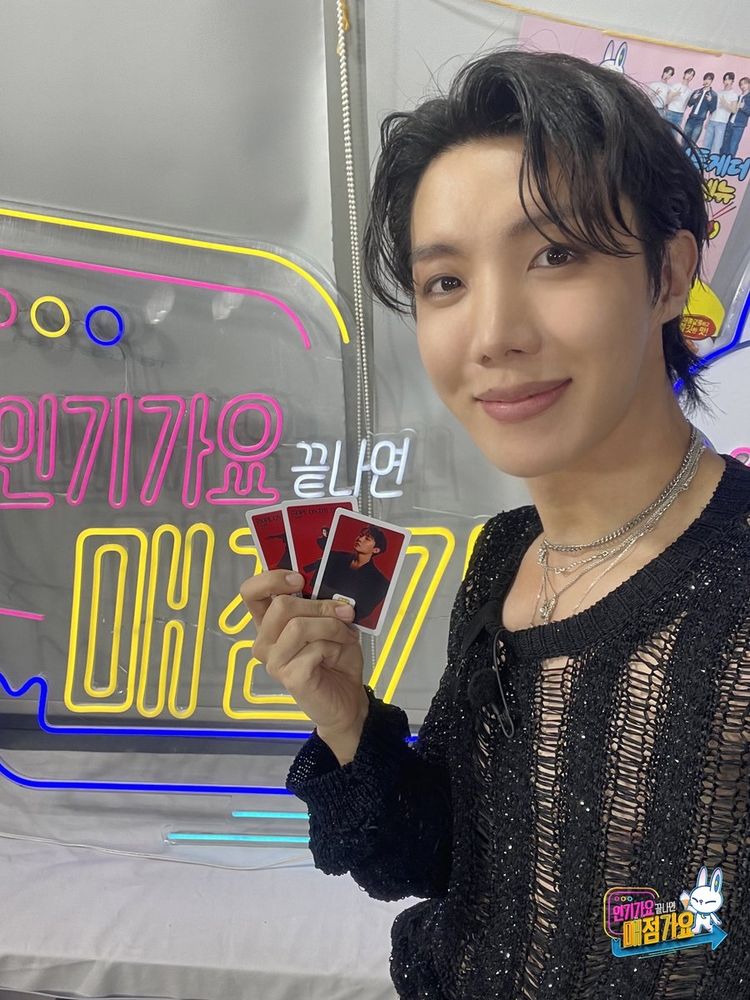 Hobi wearing a distressed black sweater holding three little cards and smiling for the camera.