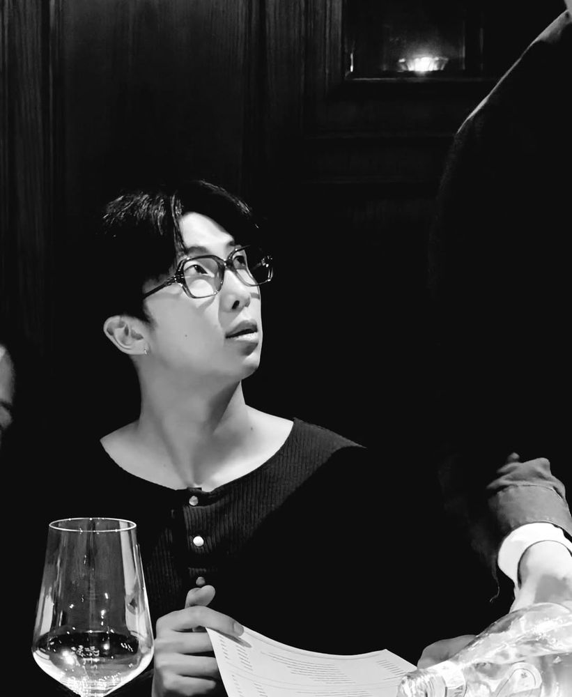 Black and white photo of Namjoon sitting at a table in a restaurant. I. Front of hin is a glass of wine. 