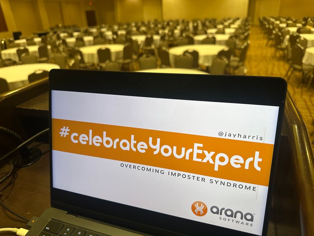 An open laptop on a podium overlooking a large empty ballroom of tables. The laptop is displaying a title slide of “Celebrate your Expert” by Jay Harris.