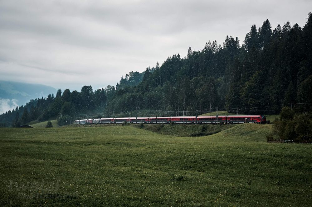 Ein Railjet fährt am Waldrand in einer Kurve entlang.. Es regnet sehr stark.