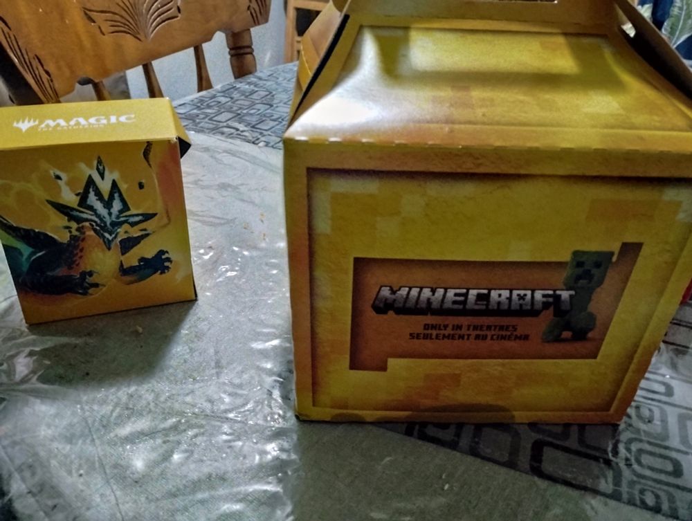 Image of two yellow boxes on a grey table with dark grey table cloths. On the left, a smaller yellow Magic the Gathering box is shown with a green and yellow dragon on it. The dragon has four spiky green and white horns, two in the middle and one on each side of the dragon's face.

The larger box has the text "Minecraft: Only in Theatres, Seulement au cinéma: It over a darker yellow text box. A Creeper, a green block creature with two square eyes, a black frowning mouth and four legs looks at the camera. 