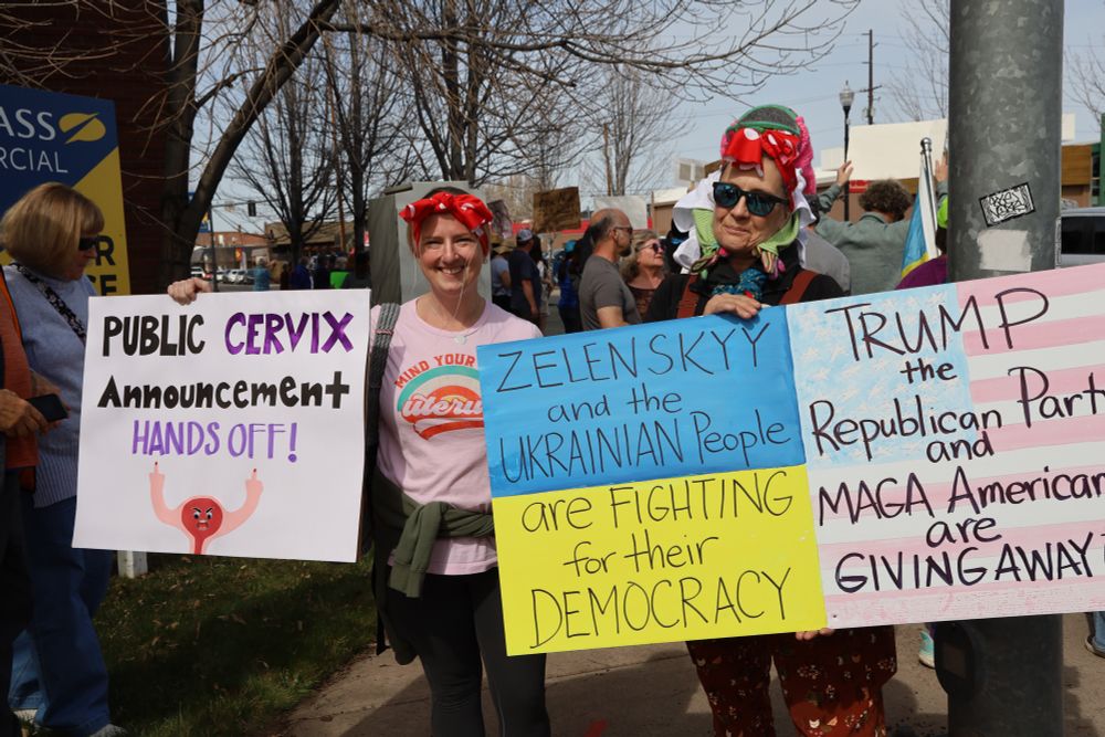 Two smiling protesters holding colorful signs. One sign humorously reads “Public Cervix Announcement HANDS OFF!” accompanied by a cartoon uterus, while the other sign is in support of Ukraine, reading “Zelenskyy and the Ukrainian People are FIGHTING for their DEMOCRACY” and criticizing Trump, the Republican Party, and MAGA Americans.
