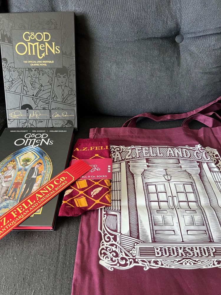 A photo of the good omens hq in a black background with some goods: a pair of Aziraphale socks, a tote bag from his bookshop and a leather bookmark 🔖 