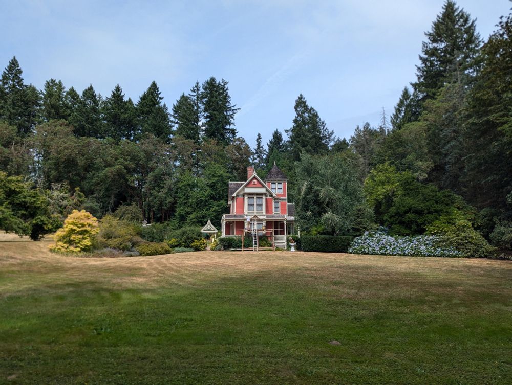 A narrow three-story red Victorian house with a turret surrounded by tall pine trees with a large grassy lawn