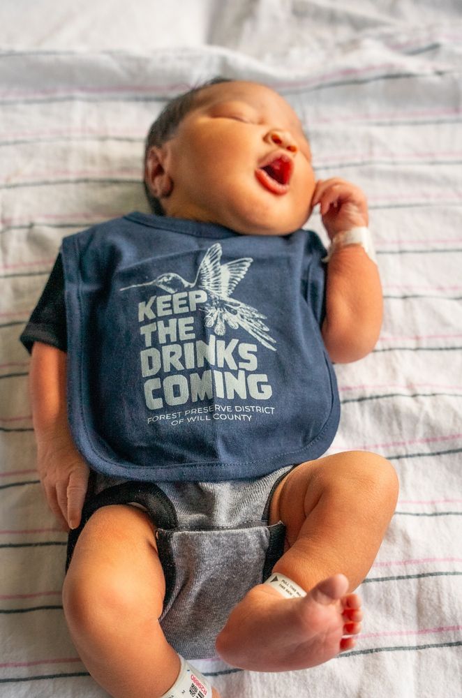 Baby wearing a bib that says keep the drinks coming, featuring a hummingbird.
