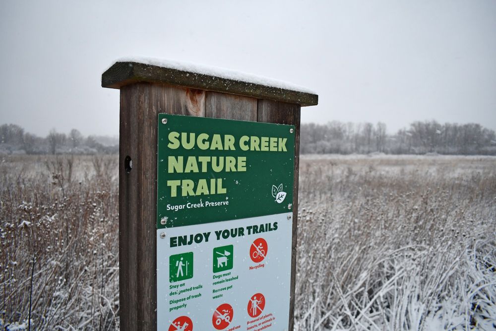 A trailhead sign with snow on top with a view of a prairie in the background.