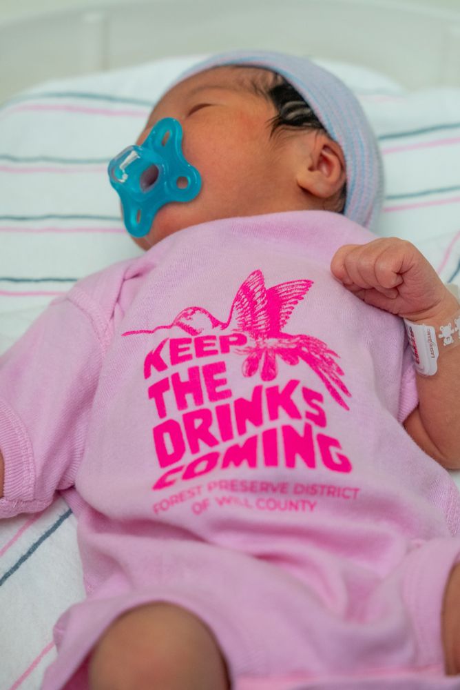 Baby wearing a pink onesie that has a hummingbird on it and says keep the drinks coming. 