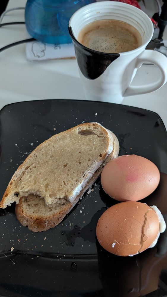 Coffee, sourdough bread with a bite out of it and 2 eggs sitting on a black plate for breakfast