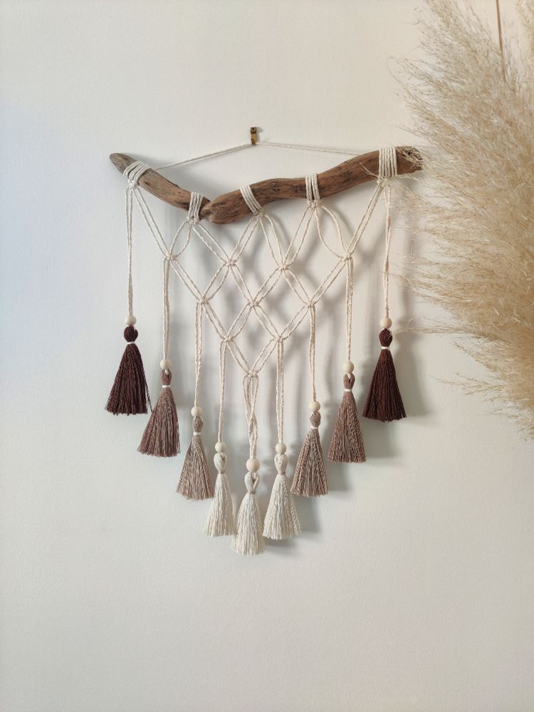 Tenture en macramé accrochée à un mur blanc. Elle est composée de noeuds plats formant un triangle avec des pompons de couleurs marrons et beige au bout.