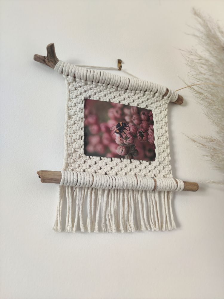 Porte photo en macramé. Deux morceaux de bois flotté sont parallèles et rejoint par un tissage de nœuds plats en corde blanche. Une photo est accrochée elle représente une abeille en train de butiner des fleurs roses.
