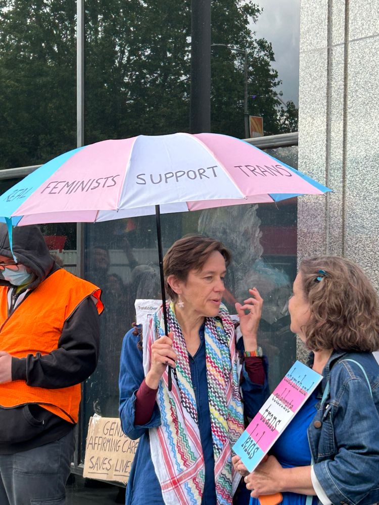 Two older supporters carry signs & umbrellas in trans flag colors. One message reads “Real Feminists Support Trans Rights”