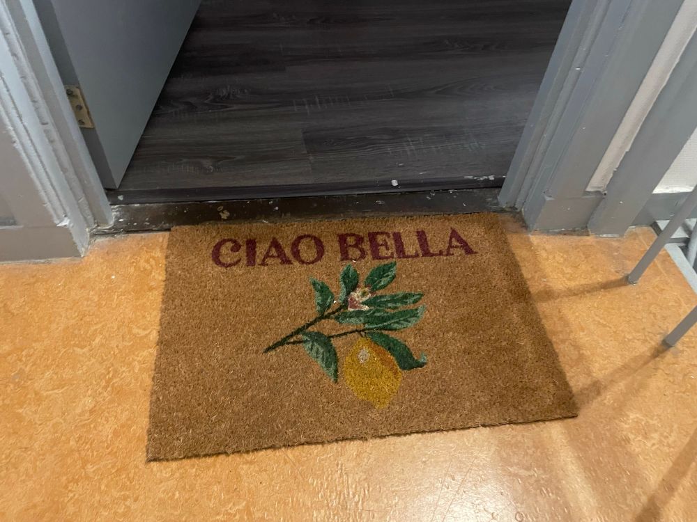 Deurmat gemaakt van kokosvezel met de tekst ‘ciao bella’ en een tekening van een tak met een peer (?) erop. 