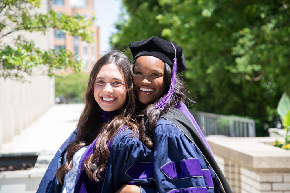 Two Georgetown Law graduates in regalia pose together, smiling and standing in the sunlight, with trees and a campus building in the background.