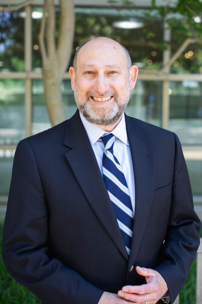 Joshua C. Teitelbaum, newly appointed Interim Dean of Georgetown Law and Executive Vice President of Georgetown University, stands smiling in a navy suit and striped tie in front of the Eric E. Hotung International Law Center Building.