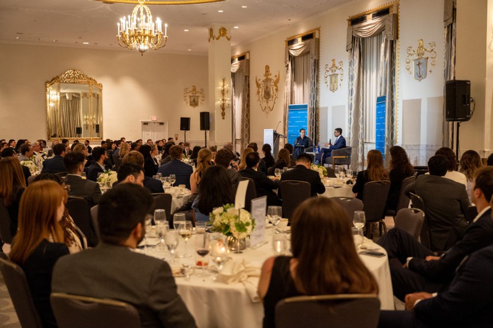 A wide view of the banquet hall at the Mayflower Hotel, showing guests seated at round tables as Judge Sri Srinivasan speaks on stage in a fireside chat with Yasmeen Rose.