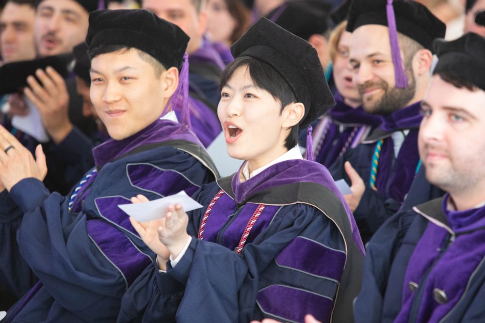 Georgetown Law graduates seated at the commencement ceremony cheer and clap while wearing academic regalia. One student in the center reacts with visible excitement.