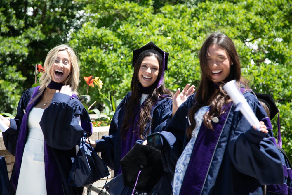 Three Georgetown Law graduates in regalia smile and celebrate outdoors, holding diplomas and graduation caps on a sunny day with greenery and flowers in the background.