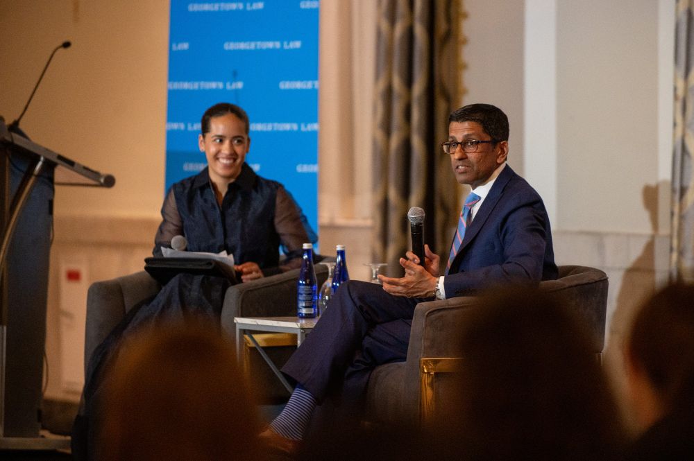 Judge Sri Srinivasan speaks into a handheld microphone during a fireside chat on stage with Yasmeen Rose, who is seated beside him and smiling.
