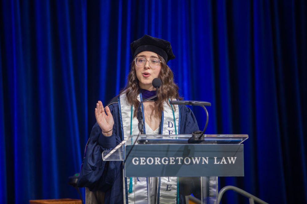 A Georgetown Law graduate speaks at a podium with the "GEORGETOWN LAW" wordmark, wearing graduation regalia and glasses. The stage backdrop is blue with spotlights.