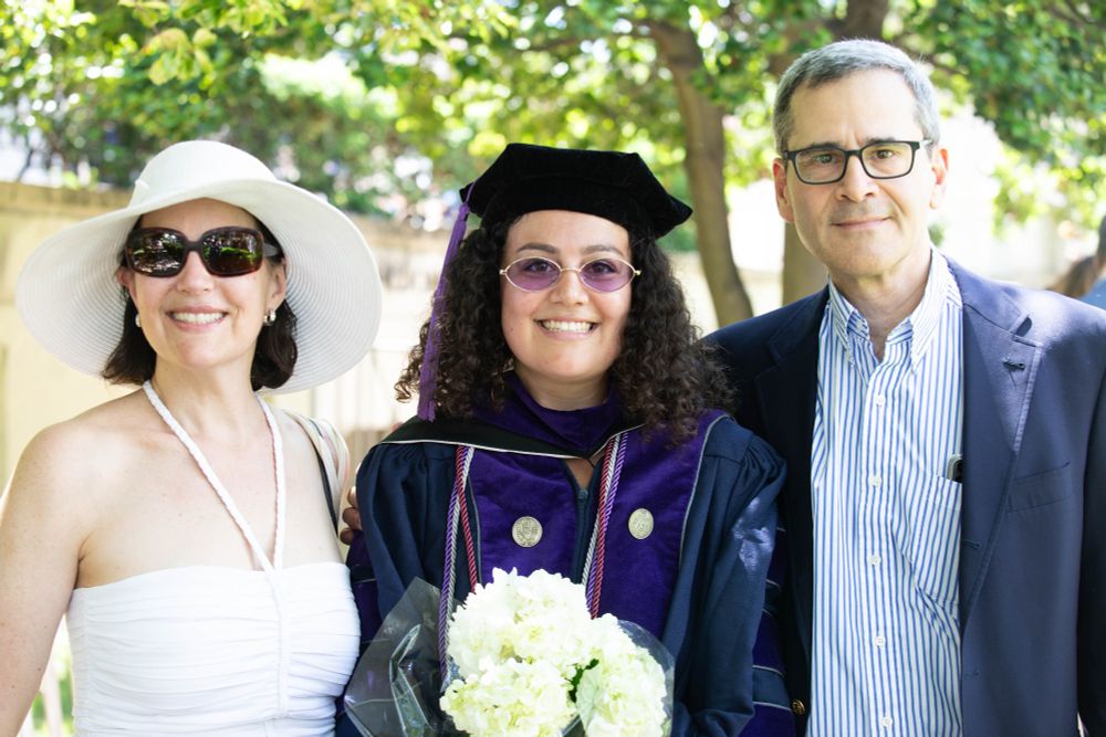 A Georgetown Law graduate in regalia stands with two loved ones, smiling and holding a bouquet of white flowers. They are outdoors under the shade of leafy trees.
