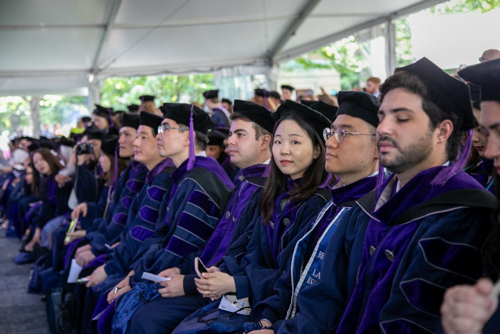 A large group of Georgetown Law graduates sits under a commencement tent, dressed in purple gowns and caps, attentively watching the ceremony.