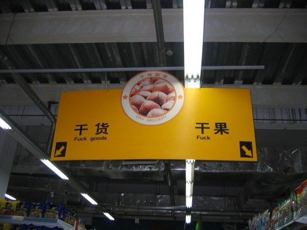 Sign over a grocery aisle. It's a big yellow sign with text on each side and arrows pointing to the side of the aisle for the text. On one side in Chinese it says 干货, which means "dried food" but is translated in English as "Fuck goods" and on the other side in Chinese it says 干果, which means "dried fruit," but is translated in English as "Fuck."