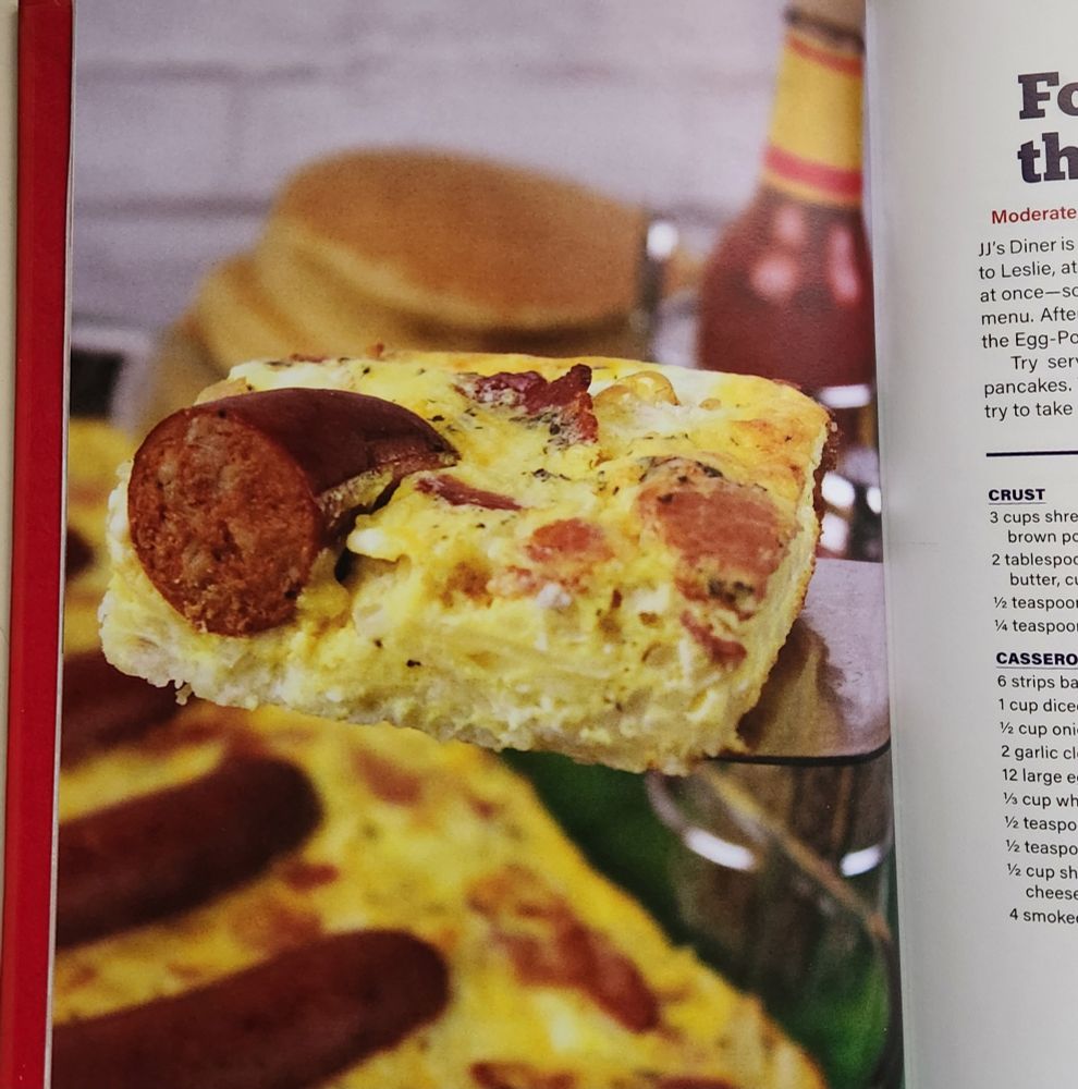 Photo of Four Horse Meals of the Egg-Pork-alypse. A slice of egg casserole studded with various meats and huge chunk of sausage is being lifted on a serving tool. In the background slightly out of focus is a stack of pancakes and bottle of hot sauce.