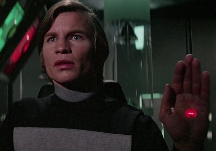 A scene from the film Logan's Run. Michael York (Logan) in a black and grey uniform raising his hand, showing a blinking red crystal embedded in the palm.
