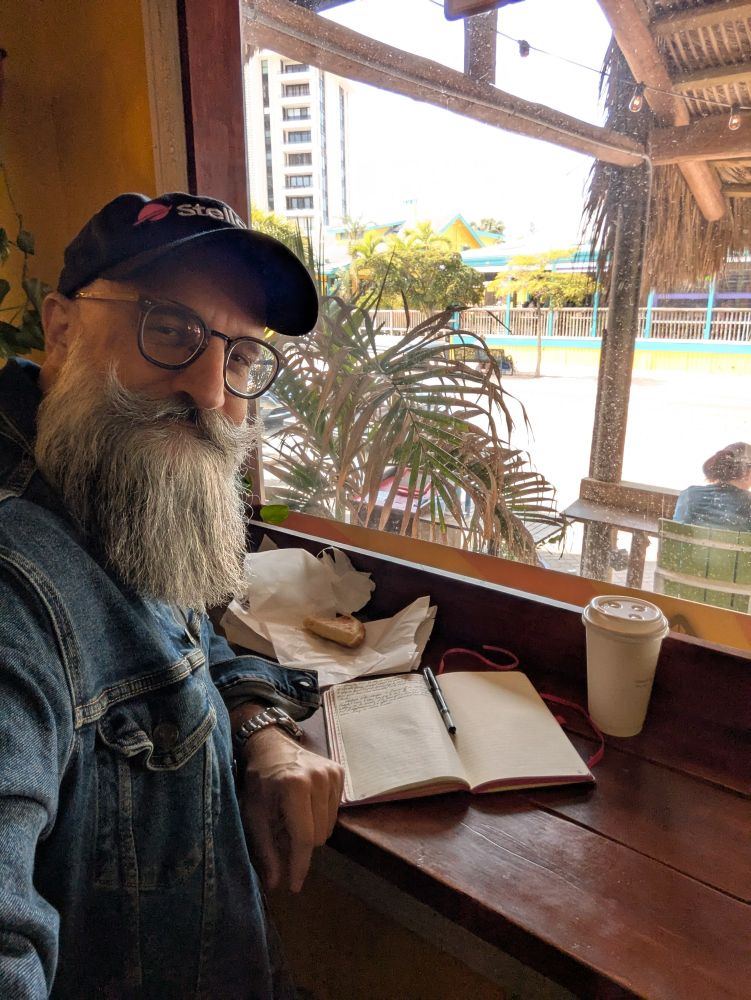 Bearded guy sitting at a counter with a journal, a pen, a half-eaten empanada, and a cup of coffee. The view from the barstool through a picture window shows a tropical street scene on a very sunny day 