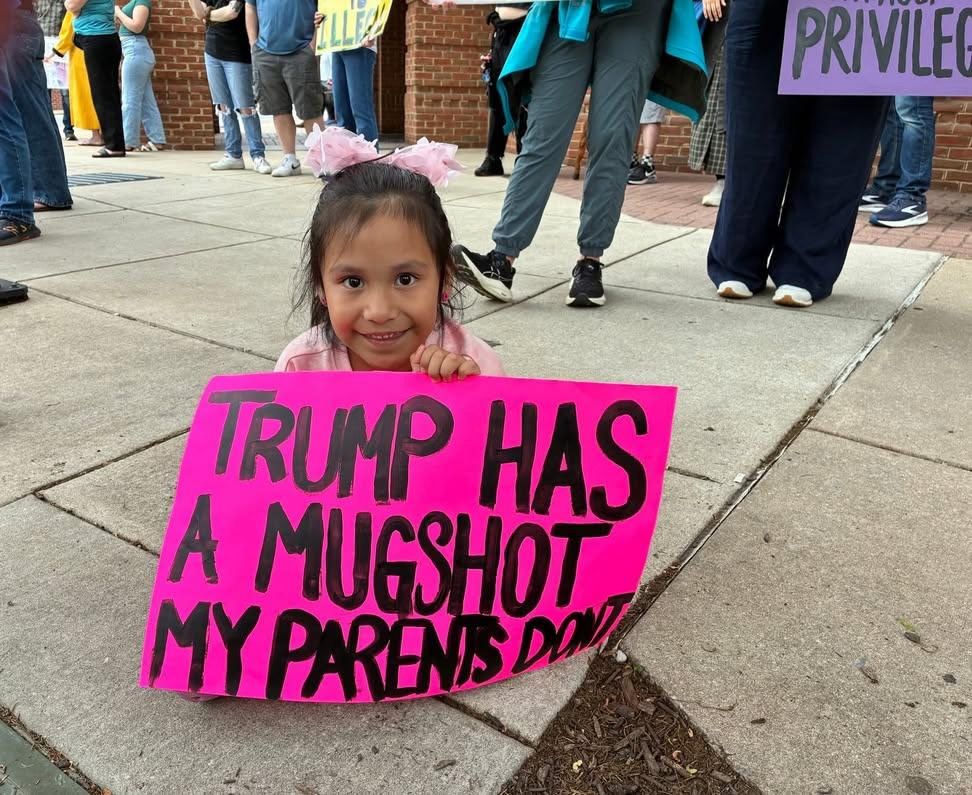 A little girl holding a pink protest sign with black writing that says TRUMP HAS A MUGSHOT MY PARENTS DONT