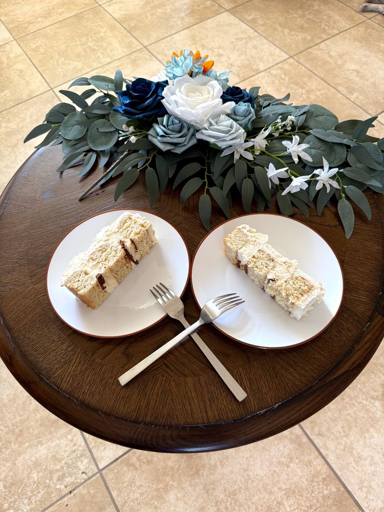 Round dark wood with grain small tabletop with 2 white round plates each with slice of wedding cake angled in towards center of table. Two forks tines down on plates handles crossed
 Top of table is spray of drak green Greenway with white and various shades of blue flower accents.