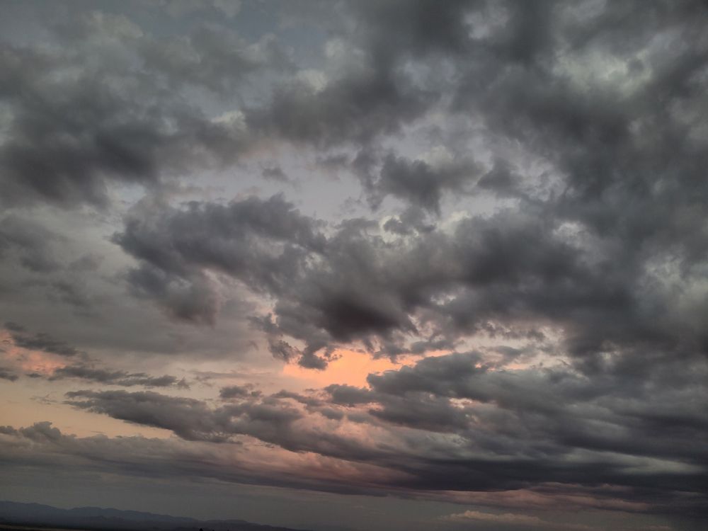 Picture is dominated by darker grey clouds. A few pockets of peachy-orange sky and under lit clouds show in the distance. Bare bottom of image is grey silhouette of mountains in the distance.