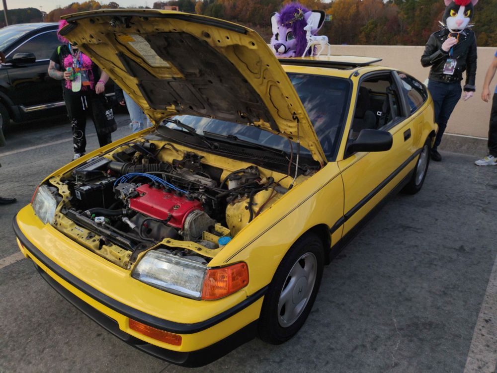 Yellow Honda CRX hatchback with the hood open. The car is a two door, a hatchback, a sunroof, and an engine with a red cover. The car is pristine.