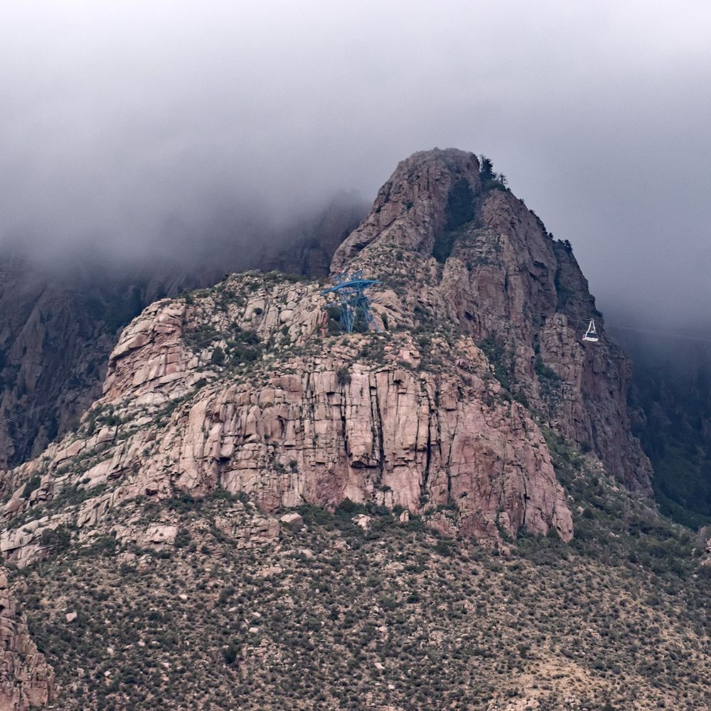 An enormous granite crag dominates the image. Perched on a ledge on the crag, a blue tower for an aerial tramway looks tiny. To the right of center, a passenger gondola looks equally small. The upper third of the image is dominated by gloomy clouds.
