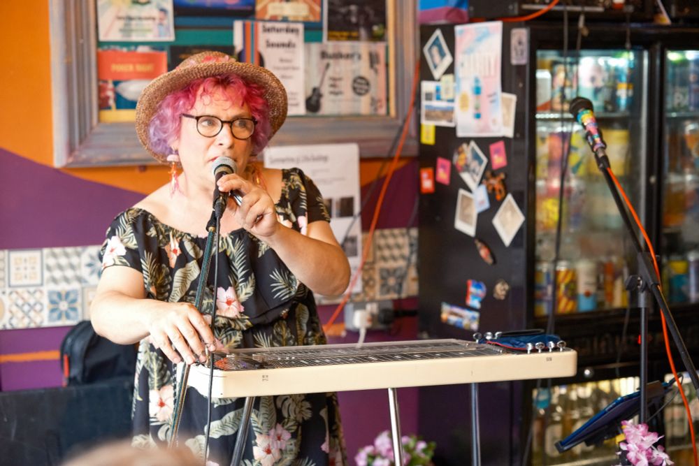 A person in a flowery dress and straw hat is talking into a microphone at a very small pub in between playing the Hawaiian Guitar. 